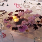 Fehér terítőn egy üveg tálon, rózsaszín és bordó virágok és teamécsesek.