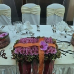 Egy fehér hosszú asztalon, téglatest alakú csokor, szélein zöld és közepén lila virágokkal.