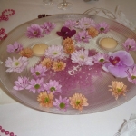 Fehér terítőn egy üveg tálon, rózsaszín és narancs virágok és teamécsesek.