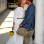 A vőlegény fehér oszlopnak támaszkodva, a menyasszony szembe állva megcsókolja, kezében egy sárga kis csokorral.