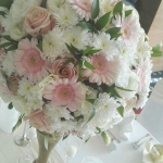 Felülről egy fehér, rózsaszín csokor, réz vázában, fehér terítős asztalon.