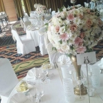Fehér terítős asztalon, magas réz vázban, fehér és rózsaszín virágok.
