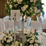 Fehér terítős asztalon, magas ezüst vázában fehér virágos csokor, mellette fehér virágos csokrok.