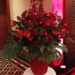 Fehér terítős kis asztalon, piros vázában piros virágokból álló csokor.