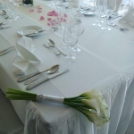 Hosszú asztal fehér terítővel, fehér étkészlettel, poharakkal és a bal alsó sarokban egy fehér kis virágcsokorral.