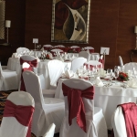 Fehér körasztalok, bordó keskeny terítővel, közepükön vörös rózsás csokrokkal, a háttérben modern festménnyel.