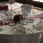 Fehér körasztal, bordó keskeny terítővel, közepük vörös rózsás csokorral.