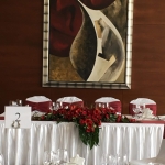 Az ifjú pár fehér asztala, bordó keskeny terítővel. A falon mögöttük egy nagy modern festmény.