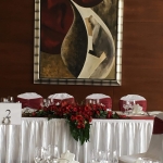 Az ifjú pár fehér asztala, bordó keskeny terítővel. A falon mögöttük egy nagy modern festmény.