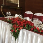 Egy esküvői asztal fehér és bordó terítővel, egy hosszú vörös rózsákból álló csokorral.