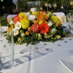 Egy fehér körasztal közelről. Rajta poharak vízzel és egy sárga, narancs és fehér virágcsokorral a közepén.