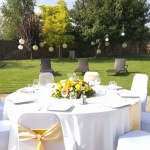 Nagy kertben barna székek, hátul barna kerítés és fák. Hozzánk közel fehér körasztal, közepén sárgás csokorral és fehér székekkel.