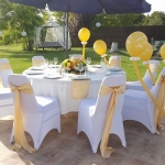 Egy kertben fehér körasztal, fehér székekkel, sárga lufikkal, kék nagy napernyővel.