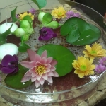 Egy kör alakú üvegtálban víz, és a vízen sárga és lila virágok és nagy zöld levelek.