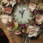 Egy fonott széken lóg egy óra ami körül fehér és rózsaszín virágokból van koszorú készítve.