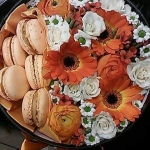 Egy kör alakú virágtartóban fehér és narancs színű virágok és rózsaszínű hamburger alakú dekorációk.