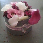 Egy henger alakú széles virágtartóban fehér, lila és rózsaszínű virágok.