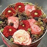 Egy henger alakú széles virágtartóban rózsaszínű, piros virágok és bogyós virágok.