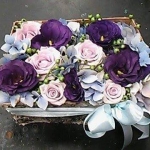 Egy tégla alakú, fa láda, lila, rózsaszínű és fehér virágokkal, fehér masnival.