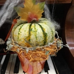 Zongora billentyűin egy piros kaspóban fehér, zöld tök, őszi falevéllel és pókhálóval.